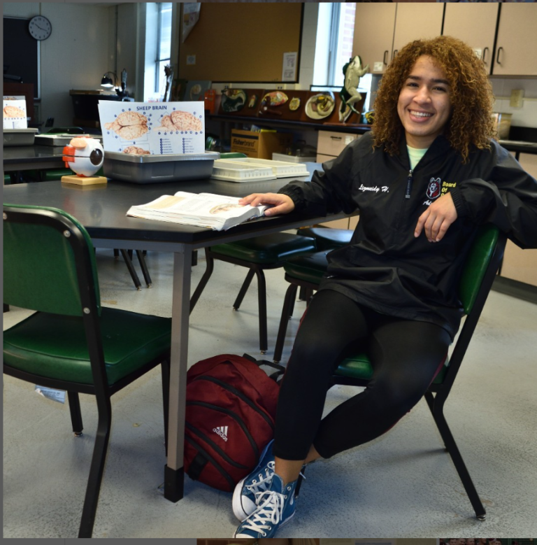 Lizmeidy Hernandez '20 is wearing a black athletic zip hoodie and black leggings with blue converse sneakers and sitting at a table in a computer lab with several anatomy models.