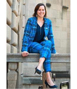 PA State Representative, Emily Kinkead sits atop a marble railing against a marble backdrop, suggesting a political building. She is wearing a blue and black plaid blazer and pants with a black top.