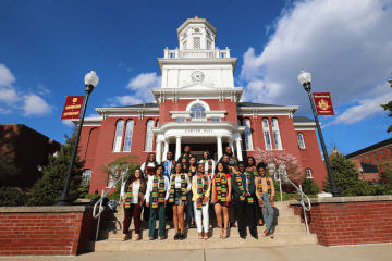 A group of new graduates stand on the steps of Carver Hall for a group photo following their Frist World ceremony