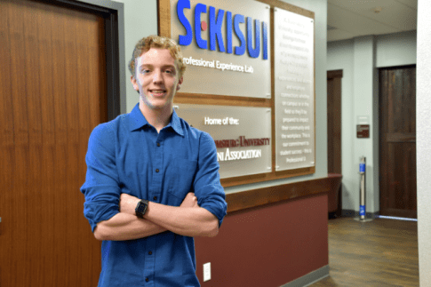 Robert Bozarth '23 stands next to the SEKISUI sign in the hallway of the Alumni and Professional Engagement space on the third floor of the Greenly Center in downtown Bloomsburg.