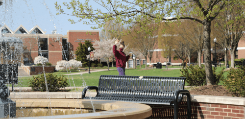 Student waving while walking in front of the fountain