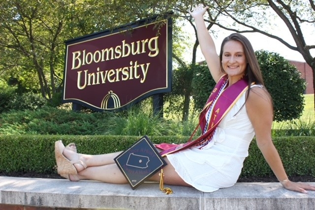 Angela DiPasquale with her graduation stole and cap sits atop the brick entrance to Bloomsburg University