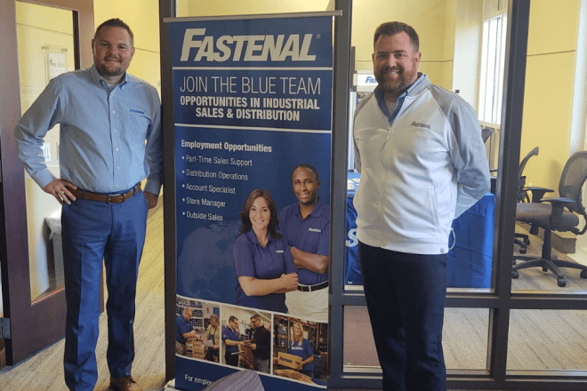 Two Fastenal employees stand on either side of a tall blue roll-up banner depicting the company logo and two smiling employees